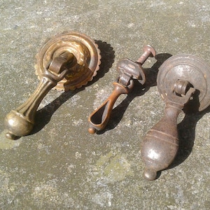May include: Three antique brass drawer pulls in varying designs. The pulls have ornate details and are in shades of gold and bronze. The drawer pulls are likely for furniture restoration or vintage decor.