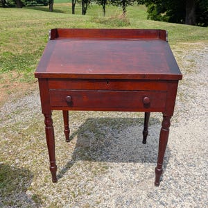 May include: A dark reddish-brown wooden slant-top desk featuring a hinged lid, a single horizontal drawer with two round wooden knobs, and four turned legs. The desk has a small back gallery and a keyhole centered on the front drawer panel.