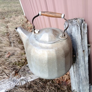 May include: A vintage metal kettle with a wooden handle and a spout. The kettle has a rounded body with a lid and a small white knob. The handle is made of wood and metal. The kettle is sitting on a wooden fence.