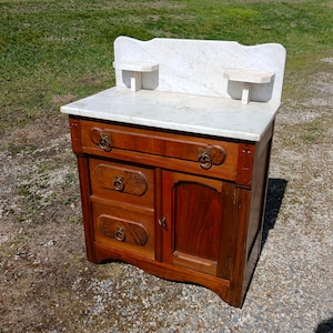 May include: Antique wooden washstand with a marble top and backsplash. The washstand features two drawers, a cabinet door, and brass ring pulls. Two small marble shelves are attached to the backsplash. The wood has a rich, warm tone.