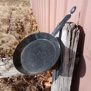 May include: A black speckled frying pan with a long, flat handle. The pan is round with a slightly raised edge. The handle has a loop at the end for hanging. The pan is resting on a weathered wooden post.