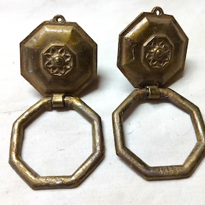 May include: Two antique brass drawer pulls. Each pull features an octagonal backplate with a floral center and a hinged, octagonal handle. The brass has a tarnished, aged appearance, suggesting vintage hardware.