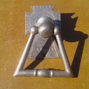 May include: A silver-toned metal door knocker featuring a triangular swinging handle attached to a decorative backplate. The backplate has a cross-like top section with circular detailing and the letters AABNA engraved in the center of the triangular base.