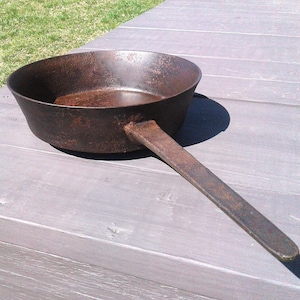 May include: A well-worn, cast iron skillet with a long, flat handle. The pan has a deep bowl and a dark, rusty brown color, suggesting age and use. The skillet sits on a weathered wooden surface, with a grassy background.