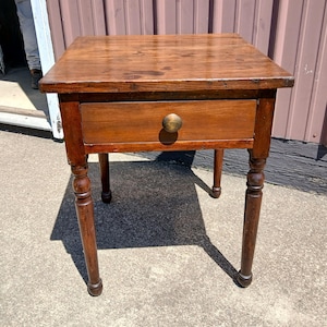 May include: A square wooden side table with a medium brown finish, featuring a single drawer with a round wooden knob. The table is supported by four turned legs with a smooth, cylindrical design, showcasing a visible wood grain texture throughout.
