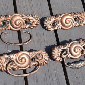 May include: Four copper-toned metal drawer pulls featuring an ornate, swirling central design flanked by leaf-like patterns. Two of the pulls include a curved, hinged handle attached to the decorative backplate, while two backplates are shown without handles.
