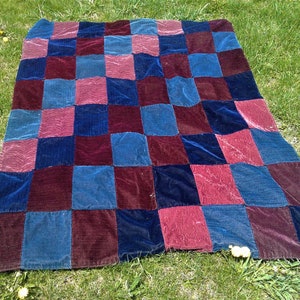 May include: A patchwork quilt with a checkerboard pattern. The quilt features squares of varying shades of blue and burgundy corduroy fabric. The quilt is lying on a bed of green grass.