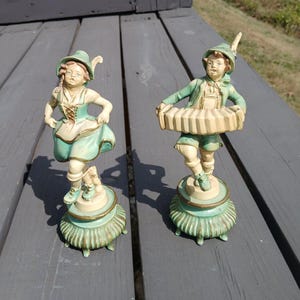 May include: Two vintage ceramic figurines of children in traditional Bavarian outfits. One figure is dancing, and the other is playing an accordion. The figurines are painted in shades of green, cream, and brown, and are mounted on decorative bases.
