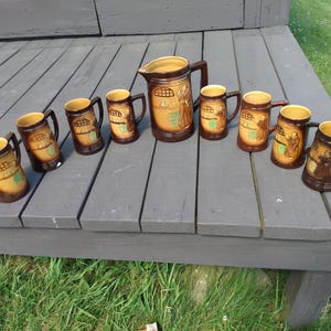 May include: A ceramic beverage set consisting of one large pitcher and seven matching mugs. Each piece features a two-tone brown and tan glaze with a decorative relief illustration of a monk figure and a window arch design on the exterior surface.