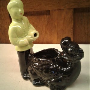 May include: A vintage ceramic planter featuring a figure in a light green outfit holding a vessel, paired with a black, stylized animal-shaped container. The planter has a glossy finish and is likely a decorative item.