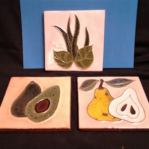 May include: Three square ceramic tiles featuring raised relief designs. One tile displays a green plant with leaves, another shows a whole and halved avocado in shades of green and brown, and the third depicts a yellow pear and a white pear slice.