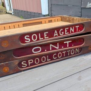 May include: A stack of three rectangular wooden display trays featuring dark brown wood frames and red glass inserts. Each tray displays white lettering that reads SOLE AGENT, O.N.T., and SPOOL COTTON. The trays have internal wooden dividers.