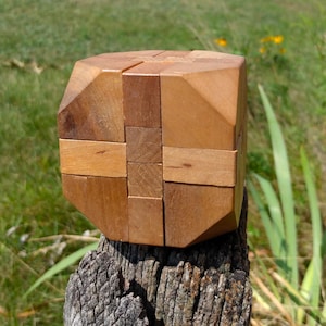 May include: A wooden puzzle cube constructed from multiple interlocking light brown pieces. The object features a geometric design with beveled edges and a smooth, natural wood grain finish, showcasing a complex assembly of rectangular and square segments.