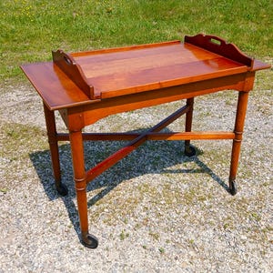May include: A wooden serving table with a removable tray top featuring cutout handles. The table has four straight legs connected by an X-shaped stretcher and is supported by four small black casters for mobility. The wood has a smooth finish.