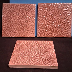 May include: Three square, dusty rose-colored tiles with a raised floral pattern. Each tile features a repeating design of stylized flowers with swirling petals and a central dot. The tiles are arranged on a dark surface with a blue background.