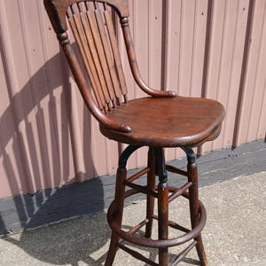 May include: A tall wooden bar stool with a dark brown finish, featuring a curved backrest with vertical slats, a contoured seat, a swivel mechanism, and a circular footrest connecting four straight legs with a central support structure.