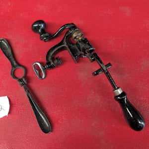 May include: A vintage black metal hand-cranked can opener with a clamp mechanism and a separate black metal handle tool. A small white paper tag with the handwritten text Lot 2 is positioned next to the metal tools on a red surface.