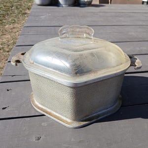 May include: A square-shaped metal casserole dish with a textured silver-tone exterior and two side handles. The dish includes a clear, transparent glass lid featuring a subtle etched geometric pattern and a central handle on the top.