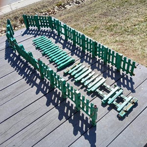 May include: A collection of small, weathered green wooden fences and walkways. The fences have a picket design, and the walkways are made of slats. The pieces are arranged on a gray wooden surface, casting long shadows.