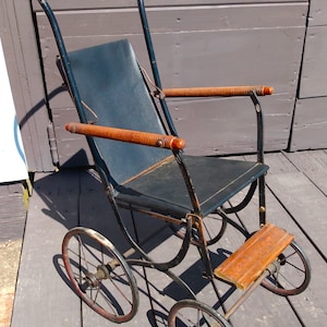 May include: An antique black metal baby carriage with a black seat and wooden armrests. The carriage has four spoked wheels with red accents and a wooden footrest. The handle is curved and made of black metal.