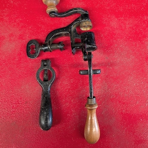 May include: A vintage black metal hand-cranked kitchen tool with wooden handles and a clamp mechanism, accompanied by two separate metal attachments. A small white paper tag with the handwritten text Lot 3 is positioned above the main device.