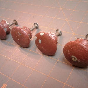 May include: Four antique cabinet knobs with a distressed, reddish-brown finish. Each knob has a rounded, oval shape and a metal screw for attachment. Some knobs show small areas of paint loss.