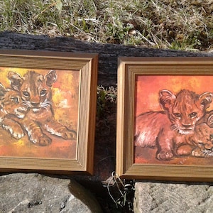 May include: Two rectangular framed paintings featuring lion cubs. The left painting shows two cubs resting against a warm orange and yellow background. The right painting depicts two cubs in a similar pose with a deep orange and red background.