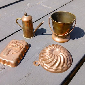 May include: Four antique copper kitchen items: a small watering can, a miniature bucket with a handle, a rectangular mold with a floral design, and a round, fluted mold. The items are arranged on a weathered gray wooden surface.