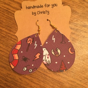 May include: Purple dangle earrings with a gold hook. The earrings feature a variety of whimsical designs including owls, lightning bolts, and a red and white scarf. The text "handmade for you by Christy" is on the brown card behind the earrings.