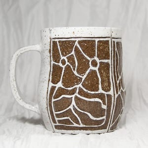 May include: A ceramic mug with a speckled white glaze and a brown textured panel featuring white hand-drawn floral and abstract line patterns. The mug has a curved handle and a cylindrical body with a flat base, showcasing a contrast between the smooth white speckled surface and the dark brown earthy clay texture.