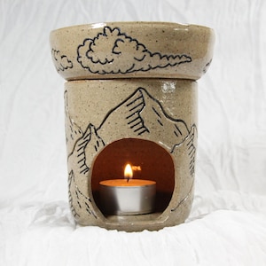 May include: A speckled beige ceramic wax warmer featuring a hand-drawn black mountain range and cloud design. The cylindrical base has a circular opening containing a lit tea light candle, while the top bowl holds wax for melting.