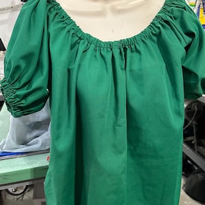 May include: A solid green blouse featuring a wide, elasticized scoop neckline and short, puffed sleeves with gathered, elastic cuffs. The garment is displayed on a mannequin, showcasing a loose, flowing fit with a simple, unadorned design.