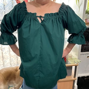 May include: A dark green peasant top with a square neckline and ruffled sleeves. The top features a tie detail at the center of the neckline. The sleeves are three-quarter length with ruffled cuffs. The top is made of a lightweight fabric.