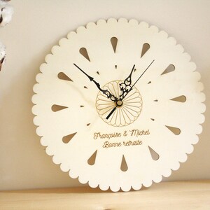 May include: White wooden clock with a scalloped edge and black hands. The clock face has a floral design and the text "Françoise & Michel Bonne retraite".