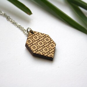 May include: A wooden pendant necklace with a geometric pattern. The pendant is a light brown color with a dark brown geometric pattern. The necklace is made of a silver chain.