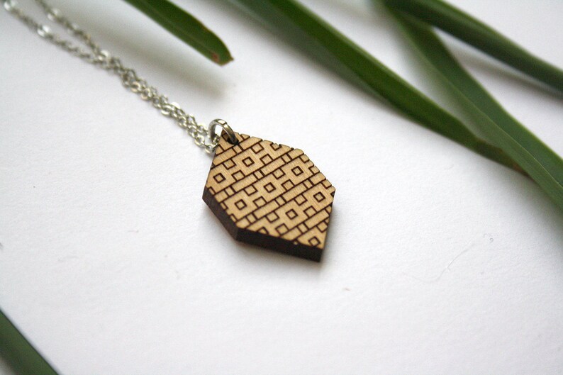 May include: A wooden pendant necklace with a geometric pattern. The pendant is a light brown color with a dark brown geometric pattern. The necklace is made of a silver chain.