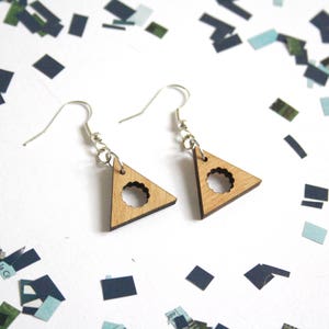 May include: A pair of wooden triangle earrings with a circle cutout in the center. The earrings are a light brown color and have silver hooks.