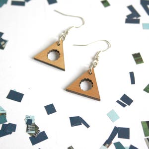 May include: A pair of wooden triangle earrings with a circular cutout design. The earrings are a light brown color and have silver hooks.