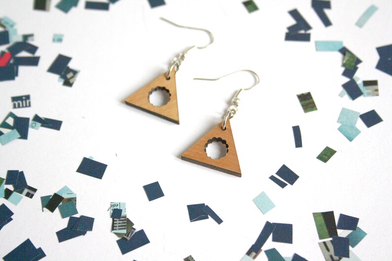 May include: A pair of wooden triangle earrings with a circular cutout design. The earrings are a light brown color and have silver hooks.