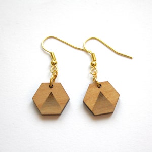 Pode incluir: Um par de brincos de ouro com pingentes hexagonais de madeira. Os pingentes têm um design geométrico com um triângulo no centro.