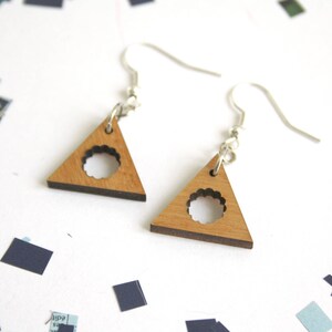 May include: A pair of wooden triangle earrings with a circle cutout. The earrings are light brown with a darker brown edge and have silver hooks.
