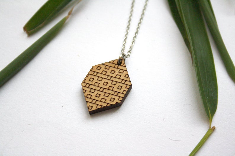 May include: A silver chain necklace with a wooden pendant. The pendant is hexagonal and has a geometric pattern carved into it.