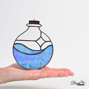 May include: Stained glass potion bottle with a black outline and a cork top. The bottle is filled with blue and purple glass, resembling liquid. The glass art is suspended by a thin wire. The bottom right corner has the text "Bright Art Glass".