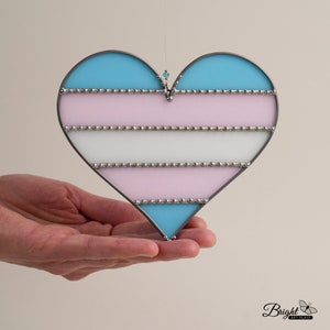 May include: A stained glass heart-shaped ornament featuring the transgender pride flag colours: light blue, pink, white, and light blue. The heart is outlined in silver and has silver beads around the edges of each colour stripe.