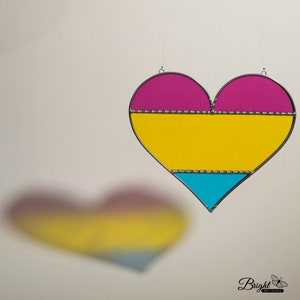 May include: A stained glass heart-shaped ornament with a pink, yellow and blue design. The ornament is hanging from a white ceiling.