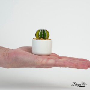 May include: A small green and yellow glass cactus in a white ceramic pot. The cactus is sitting on a hand.