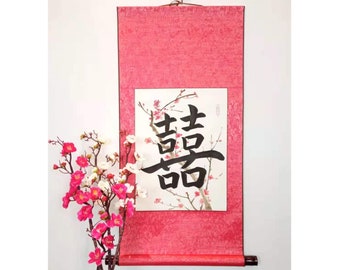 May include: A red scroll with a black Chinese calligraphy character on a white background. The character is surrounded by a painting of pink blossoms. The scroll is hanging on a white wall with a pink flower arrangement in the foreground.