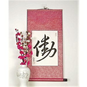 May include: A red and white scroll with black calligraphy that reads "Shou" which means longevity in Chinese. The scroll is hanging on a white wall with a pink flower arrangement in a white vase.