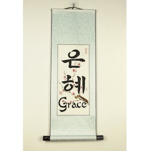 Könnte beinhalten: Eine traditionelle chinesische Kalligraphie-Rolle mit dem Wort "Gnade" in schwarzer Tinte auf weißem Hintergrund. Die Rolle ist mit einem zarten Zweig mit rosa Blüten verziert.