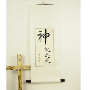 Könnte beinhalten: Eine weiße Schriftrolle mit schwarzer Kalligraphie, die "神" und "永遠愛" lautet. Die Schriftrolle ist auf einem Holzstab montiert und hängt an einer weißen Wand.